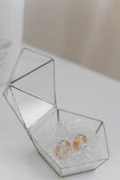 A small box with a pair of gold earrings inside. The box is made of glass and has a triangular shape. The earrings are placed on top of a bed of white beads photo