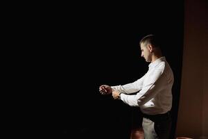 A man in a white shirt is standing in a dark room, looking at his watch. He is in a hurry and is checking the time frequently photo