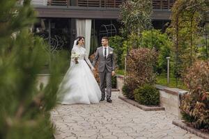 A bride and groom are walking down a path in a garden. The bride is wearing a white dress and the groom is wearing a gray suit. They are holding hands and appear to be happy photo