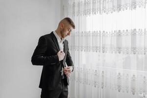 A man in a suit is standing in front of a window, adjusting his tie. Concept of formality and professionalism, as the man is dressed in a suit and tie. The window provides a view of the outside world photo