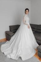 A woman in a white wedding dress stands in front of a couch. The couch is grey and has a pattern on it. The woman is wearing a veil and has her hands on her hips. The scene is intimate and romantic photo