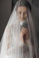 A bride is wearing a veil and holding a flower. The veil is made of a net-like material and covers her face photo