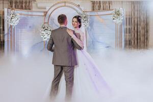 A bride and groom are dancing in front of a blue archway. The bride is wearing a white dress and the groom is wearing a gray suit. The atmosphere is romantic and joyful photo