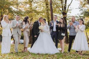 A group of people are posing for a picture, with a bride and groom in the center. The bride is wearing a white dress and the groom is wearing a suit. The group is standing in a park photo