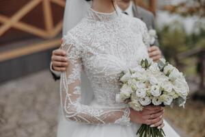 A bride and groom are posing for a picture with a bouquet of white flowers. The bride is wearing a white dress and the groom is wearing a suit. Scene is happy and celebratory photo