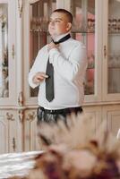 A man in a white shirt and black tie is standing in front of a cabinet. He is adjusting his tie and looking at the camera. Concept of formality and attention to detail photo