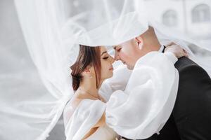A bride and groom are embracing each other under a white veil. The bride is wearing a necklace and the groom is wearing a tie. Scene is romantic and intimate photo