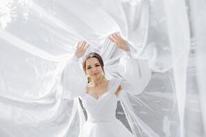 A woman in a white wedding dress is standing in front of a white curtain. She is smiling and she is happy photo
