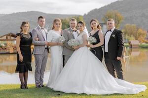 A group of people are posing for a photo, including a bride and groom. The bride is wearing a white dress and the groom is wearing a suit. The group is standing in front of a body of water photo
