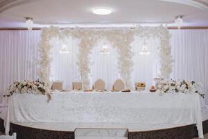 A white table with a white tablecloth and white flowers on it. The table is set for a wedding reception photo
