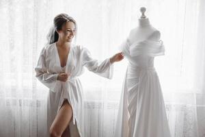 A bride is standing in front of a wedding dress, smiling photo