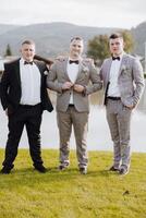 Three men in suits pose for a photo by a body of water. Scene is formal and relaxed, as the men are dressed in suits and ties, but they are also standing in a grassy area by the water
