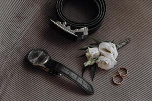 A watch, a belt, and a boutonniere are displayed on a gray surface. The watch is black and has a silver band photo