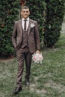 A man in a suit and tie stands in front of a bush holding a bouquet of flowers photo