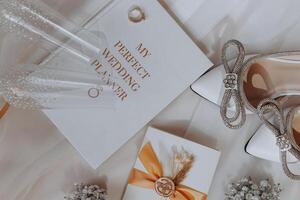 A wedding planner sits on a table with a pair of high heels and a ring. The planner and is surrounded by other items, including a glass and a vase photo