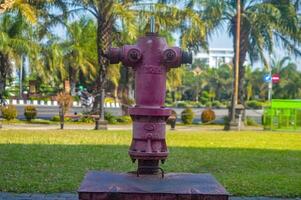 Old red fire hydrant on a grassy area with palm trees and a city street in the background, Indonesia, 27 July 2024. photo