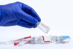 Vaccine vials and needles lie on a white background, a hand in a blue glove holding the vaccine vials above it photo