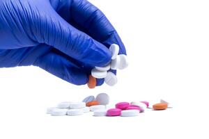 Blue gloved hand of doctor or nurse taking pills from spilled multicolored pills on white background. photo