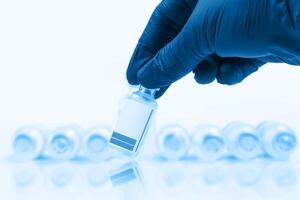 Blue gloved doctor hand holding a vaccine vial in front of a blurred row of vaccine vials on a blue background. photo