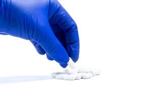 Blue gloved doctor hand holding white pill with white pills lying in group on white background. Closeup photo