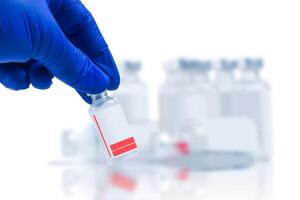Hand in blue glove holding vial with vaccine sample in front of blurred vaccine vials on white background. Closeup photo