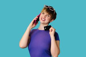 The concept of online shopping. A Caucasian young woman holds a credit card, talks on a smartphone, makes a purchase, orders delivery, standing on a blue background. photo
