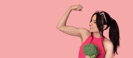 A smiling white girl with fresh broccoli in one hand and showing off her biceps in the other. Panoramic banner, free space. photo