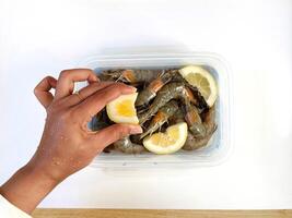 A woman hand is squeezing lemon over raw shrimp in a plastic container photo