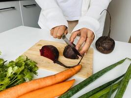 Cooking concept. Unrecognizable woman in white clothes making healthy dinner, cropped of lady preparing delicious meal at home, cutting beetroot vegetables on chopping board, kitchen interior photo