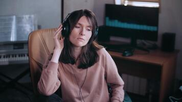 A beautiful girl with headphones listens to music in a chair at the computer and sings, smiles. Unfolds from the computer to the camera. Looks at the camera. The equalizer is playing on the monitor. video