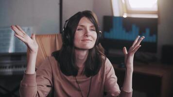 A beautiful girl with big headphones listens to music in a chair at the computer, dances, looks at the camera and smiles, the equalizer is playing on the monitor. Shakes his head to the rhythm. video