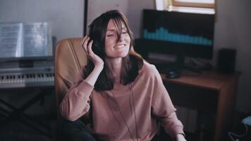 A beautiful girl with headphones listens to music in a chair at the computer. Dancing and smiling. The equalizer on the monitor. He shakes his head to the beat and taps his finger. Slow motion. video