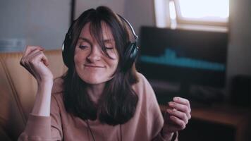 uma lindo menina com fones de ouvido escuta para música dentro uma cadeira às a computador, danças, olhares às a Câmera e sorrisos, a equalizador é jogando em a monitor. shakes dele cabeça para a ritmo. lento movimento video