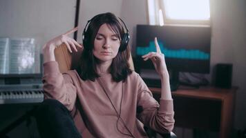 A beautiful girl with big headphones listens to music in a chair at the computer, dances, looks at the camera and smiles, the equalizer is playing on the monitor. Shakes his head to the rhythm video