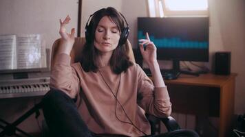 A beautiful girl with headphones listens to music in a chair at the computer, dances, looks at the camera and smiles, the equalizer is playing on the monitor. Shakes his head to the rhythm and beats video