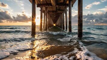 waves hitting the beach under a pier with sun coming through it, photo