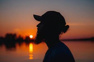 a man in sunglasses, hat and t - shirt standing by the ocean looking down on sunset horizon, photo