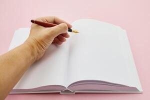 International Left-handers Day. Left hand holding a pen, writing in an open blank journal on a light pink background. photo