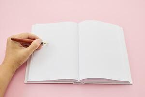 International Left-handers Day. Left hand holding a pen, writing in an open blank journal on a light pink background. photo