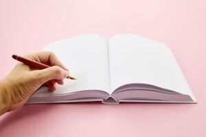 International Left-handers Day. Left Hand holding a pen, writing in an open blank journal on a light pink background. photo