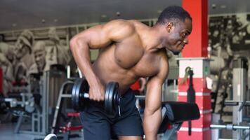 Motivated african american shirtless bodybuilder training arms and back muscles with dumbbells at modern gym. Black young man working on his biceps, lifting barbell with one hand video