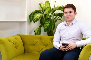Portrait of a successful man in shirt and pants, sitting on a sofa in a stylishly arranged office with a phone in his hands, conveying an image of professionalism and determination photo