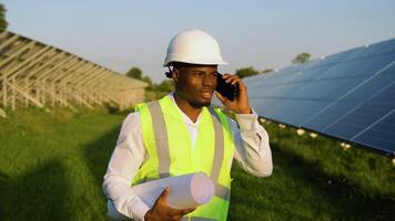 African american engineer wearing safety hard hat, holding mobile phone, making a call and walking. Engineer, worker, or architect working with Solar panel concept video