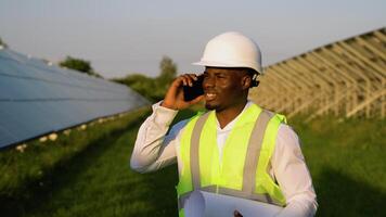 African american engineer wearing safety hard hat, holding mobile phone, making a call and walking. Engineer, worker, or architect working with Solar panel concept video