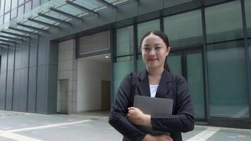 Asian businesswoman stands holding a laptop and thumbs up at the camera with a smile behind a tall building. video