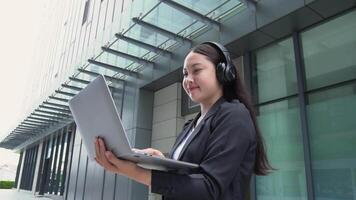 Asian business woman standing Wearing headphones using a laptop in front of a large building video