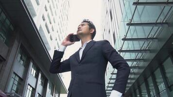 Asian businessman in black suit making phone call behind big beautiful building business investment communication concept video