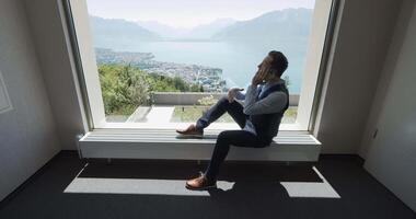 Man sitting by a window with a beautiful mountain lake view, creating a tranquil atmosphere video