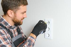 Confident professional electrician in uniform using screwdriver while replacing a socket in apartment after renovation work. photo