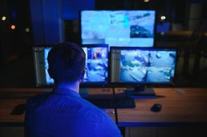 Rear view of security system operator looking at CCTV footage at desk in office photo